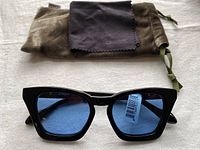 Front view of black frame Sonix Ginza sunglasses with blue lenses, showing attached price tag, with cleaning cloth and velour pouch in background.