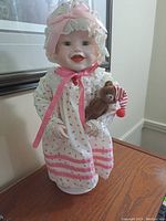 Front view of the porcelain doll Sarah wearing white bonnet and white dress with pink ribbons, holding teddy bear.