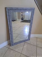 Front view of Bouclair mirror leaning against a wall on tiled floor showing grey weathered wood frame and reflective glass surface.