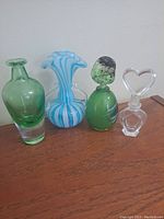 Four decorative glass items arranged on wooden surface including two green perfume bottles, one blue and white swirled vase, and one clear bottle with heart stopper