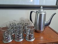 Full set of stainless steel teapot with 9 glass teacups in stainless steel holders arranged on a wooden table.