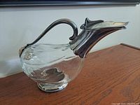 Side angled view showing clear glass body with silver plated handle and spout, on wooden surface.