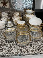 Wide view showing multiple porcelain tea cups with gold trim, glass custard cups with gold rims stacked in front, and other porcelain items in background on granite counter.