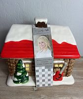 Full view of the ceramic cabin cookie jar showing the red roof, snow accents, green pine tree, and red guitar detail.