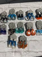 Full lot of 12 pairs of handmade earrings on display cards laid out on white cloth, featuring a mix of blue, black, orange, purple, and clear resin and glitter designs.