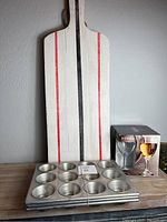 Wood charcuterie board standing vertically with handle, two boxed wine glasses, three muffin tins stacked