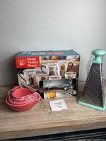 Photo showing the boxed set of 6 ceramic coffee mugs with various animal prints, ceramic flamingo shaped measuring cups nested, metal ice tongs, and metal grater with green handle