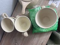 Top view of several cups and a covered casserole lid, showing creamy speckled pottery with a handle design.