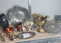 Wide view showing full assortment including brass double pour vessel, large pressed aluminum floral bowl, copper bowl, round silver-plated coasters and oval tray.
