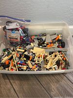 Plastic tub filled with a variety of Lego pieces in multiple colors including tan, gray, orange, and black. Includes flat plates and other brick shapes.