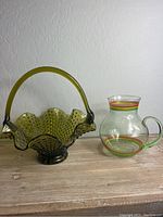Olive green hobnail glass basket with handle and green glass pitcher with painted stripes side by side on wooden surface