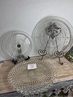 Three clear vintage glass trays displayed on a wooden surface and stands: a hobnail three-part relish tray with distinctive bubble texture, a large cubist style platter with geometric lines, and a large five-section tray with hobnail design around the edge.