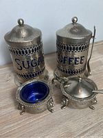 Two silver-tone metal canisters labeled 'SUGAR' and 'COFFEE' with pierced lattice design and cobalt blue glass interiors, alongside two lion's head silver-tone condiment servers with blue glass inserts and small spoons.