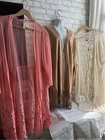 Three long cardigans hanging on hangers side by side showing styles and colors with tags visible on some.