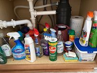 Under kitchen sink showing various house cleaning supplies, spray bottles, powder cleanser, sponges, and a plastic watering can.