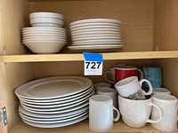 White stoneware dinner plates, lunch plates, cereal bowls, and assorted mugs stacked on kitchen cabinet shelf.