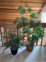 Photo showing both faux plants: a taller Schefflera plant in basket on right and shorter Fan Palm plant in blue ceramic pot on left, displayed on a table in a loft area.