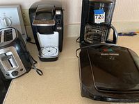 Photo showing four small kitchen appliances on a countertop: a two-slice toaster, a Keurig single-serve coffee maker, a traditional drip coffee maker, and a George Foreman grill.