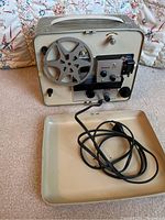 Full view of the beige and gray Kodak Brownie 310 movie projector showing one reel, attached power cord coiled inside a beige tray placed in front of the projector.