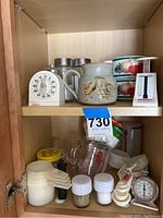 Inside view of wooden kitchen cabinet showing white mechanical kitchen timer, ceramic mugs, two salt and pepper shakers, plastic measuring cups, clear glass measuring pitcher, and white analog kitchen scale.