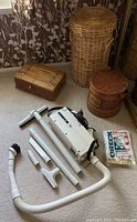 Oreck XL vacuum with multiple attachments and filter bags grouped together with a wicker hamper and two wooden boxes on carpeted floor