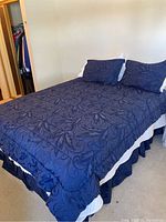 Photo shows queen bed made up with dark blue comforter and pillow shams with a leafy swirl pattern, navy blue bedskirt with white ruffle.