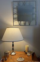 Photo showing silver base lamp with white shade next to candle, plaque, small bird figurine, and silver square vase, with mirrored wall clock hanging on wall behind.