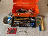 Orange toolbox with metal latches containing various tools including hammers, screwdrivers, electrical cords, fasteners, and tape, shown outside on carpet.