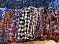 Six plaid flannel shirts in colors including red/black, purple, black/white, cream/red and red/blue checks, hanging on hangers on a bedspread.