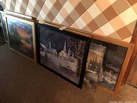 Multiple fully framed jigsaw puzzles in various sizes with wood and black frames, featuring winter and village scenes, shown leaning against a wall on a carpeted floor.