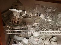 Shelf showing a ceramic ornate pitcher with plate, glass stemmed glass, cut glass bowls, and floral ceramic cups with saucers.