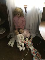 Full view of five dolls including large doll standing and smaller dolls sitting or lying on the floor in front of a curtain.