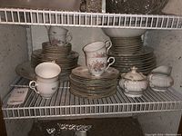 Photo showing stacked dinnerware including cups, plates, cream and sugar set on wire shelf.