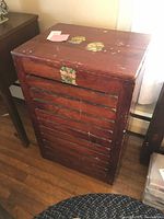 Front angle view of antique wooden quilt cabinet showing slatted front and folk art floral decoration on top and center front with visible wear and surface scratches.