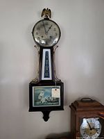 Full view of the antique banjo clock hanging on the wall showing round clock face, eagle finial, gold decorative side supports, and a painting of sailing ships in lower panel.