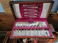 Open wooden box showing partial set of silver plated flatware with ornate handles, including forks, spoons, and knives with burgundy lining inside the case.