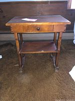 Front view of small wooden end table with drawer and lower shelf on carpeted floor.