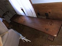 Photo of solid wood trestle bench showing rectangular top with scratches and marks, placed next to a couch and near a wooden wall