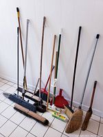 Full view of all items standing against a wall on tiled floor, shows multiple brooms, mop with sponge, dustpan, and broom heads.