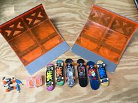All six mini fingerboard skateboards shown lined up with two orange half pipe ramps behind them, plus the small screwdriver, wheels, trucks, and motor pack seen to the left.