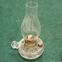 Clear vintage styled glass hurricane lamp with bulbous glass globe and rounded base, sitting upright on green carpet. Fabric wick visible inside.