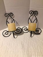 Pair of black iron wall sconces with scrollwork design and beige pillar candles placed on a fabric surface against a white background.