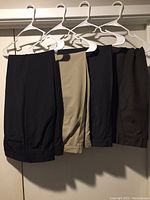Four pairs of men’s dress pants hanging on white plastic hangers, colors olive, black, gray, and tan shown.