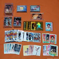 Photo showing various basketball cards spread on an orange surface including individual cards, hologram cards in plastic cases, and multiple loose cards from various eras.