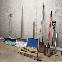 Wide shot showing all yard tools arranged against a wall, includes shovels, pitchfork, hoe, saw, clippers and hand tools