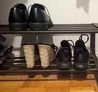 Two black IKEA PORTIS shoe racks, each holding various shoes including formal black dress shoes, black sneakers, and wedge sandals on their two slatted shelves, shown from different angles.