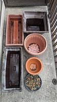 Six plastic planters and pots arranged on floor: rectangular terracotta, square beige, long black rectangular, round beige, small terracotta round with soil, small black round with soil and stones.
