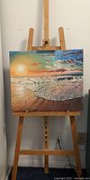 Wooden easel holding a nautical themed painted canvas depicting a beach scene at sunset with waves and clouds.