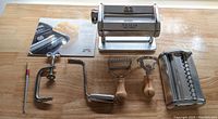 Overview of pasta machine and all accessories including manual crank, clamp, two ravioli cutters with wooden handles, and noodle cutting attachment on wood table.