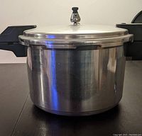Front view of aluminum pressure canner with black handles and attached lid with pressure regulator knob.